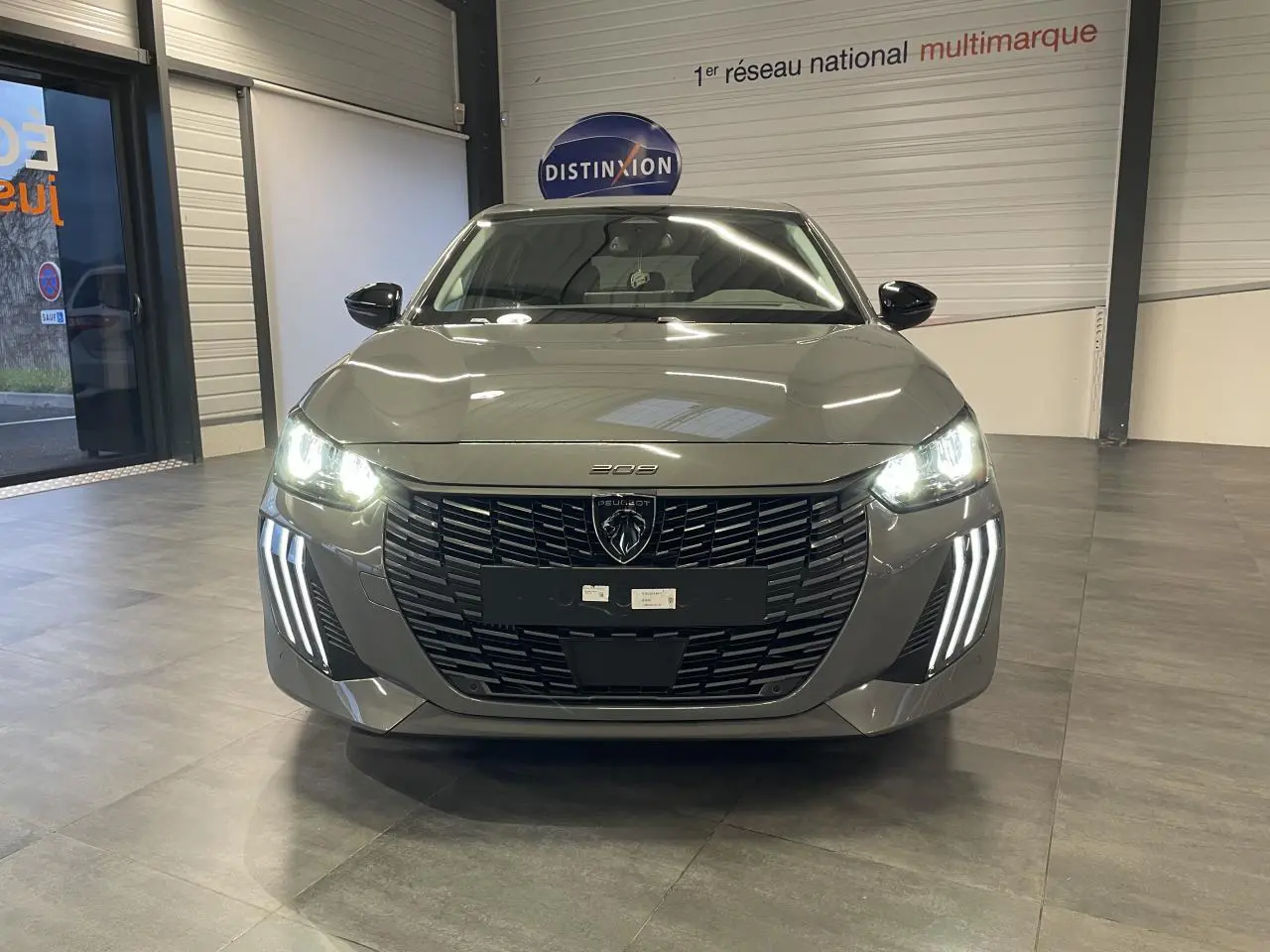 Peugeot 208 gris Selenium vue de face en intérieur, avec calandre noire et feux LED allumés.