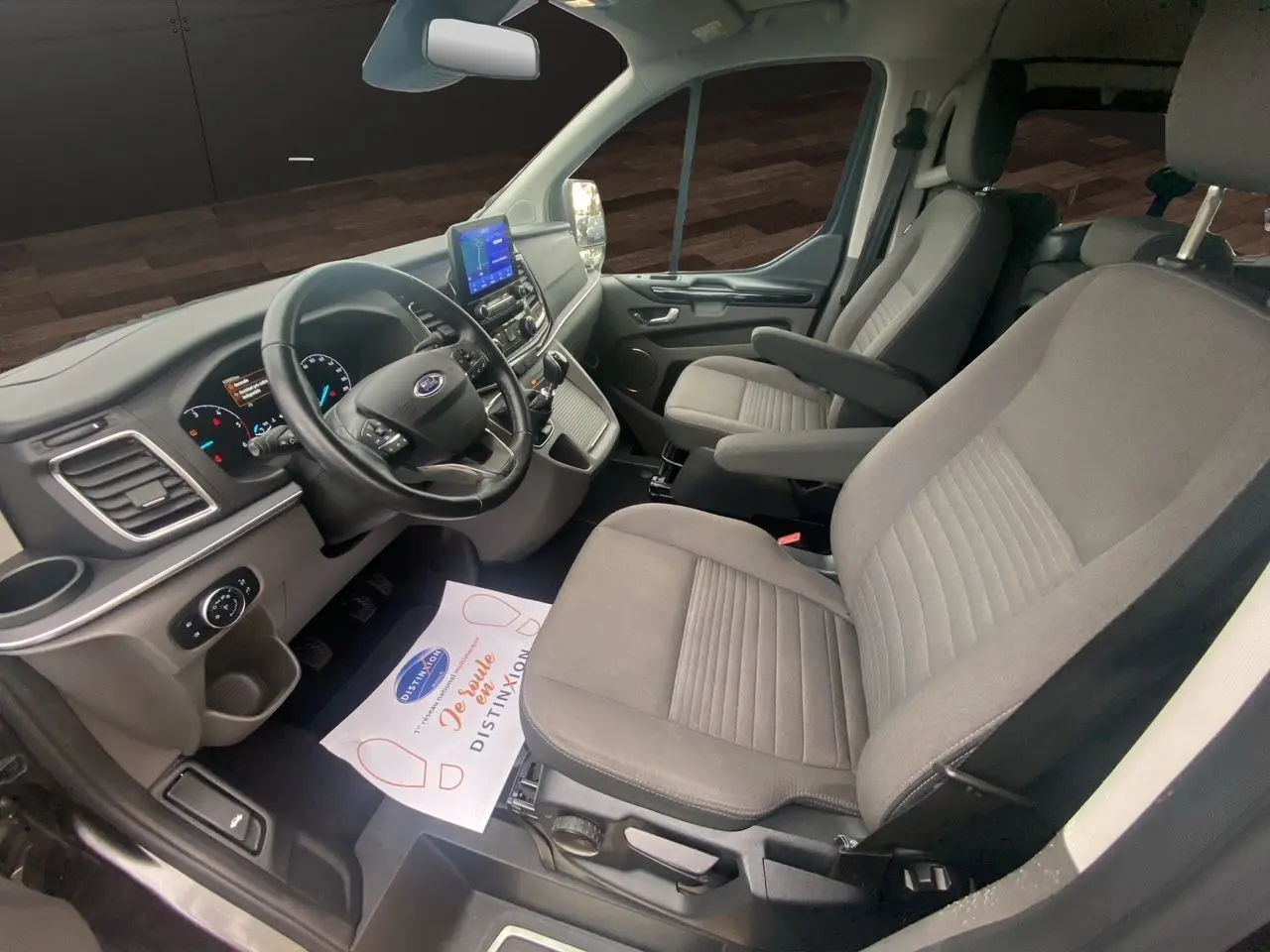 Vue intérieure côté conducteur du Ford Tourneo Custom gris magnetic, montrant le tableau de bord et les sièges avant en tissu gris.