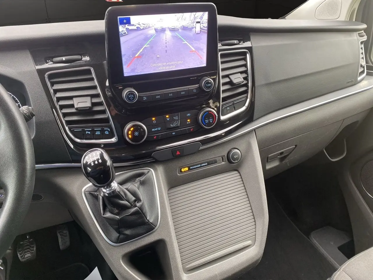 Vue intérieure du tableau de bord du Ford Tourneo Custom gris, avec écran tactile et levier de vitesses manuel visible.