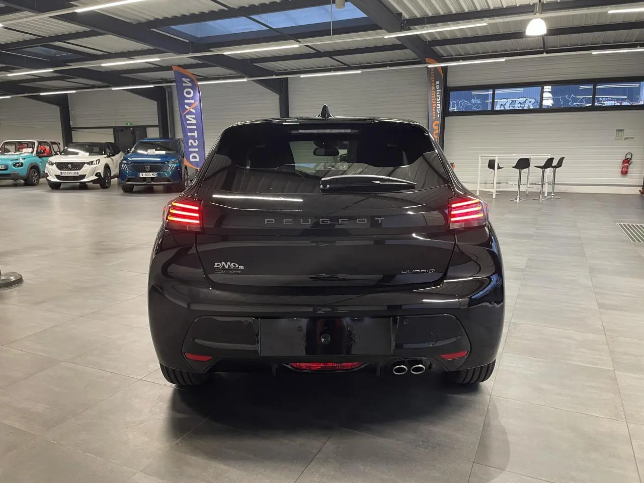 Vue arrière d'une Peugeot 208 hybride noire métallisée avec feux arrière allumés en intérieur showroom.