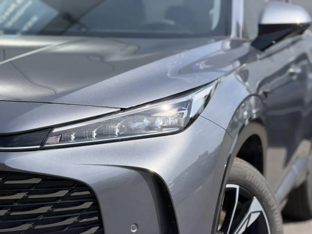 Gros plan sur l'avant droit gris métallisé du MG MOTOR EHS 1.5 Hybrid+ 224 Luxury avec phare LED et jante alliage.
