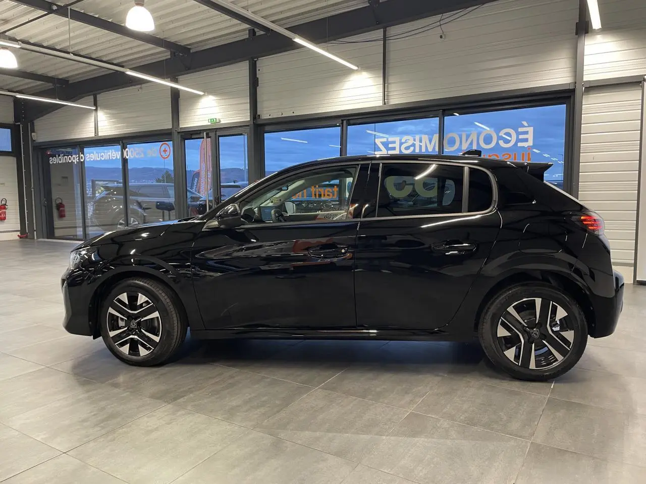 Profil droit d'une Peugeot 208 noire métallisée 2025 en intérieur, avec jantes alliage bi-tons et détails noirs brillants.