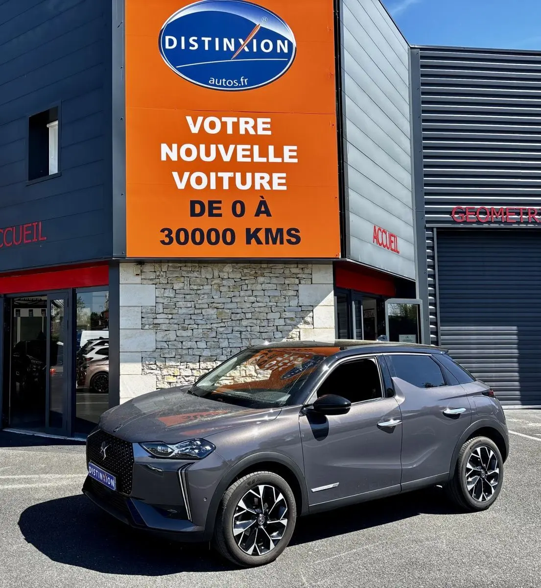 DS3 gris foncé en 3/4 avant droit, stationnée devant un bâtiment avec panneau orange Distinxion visible.
