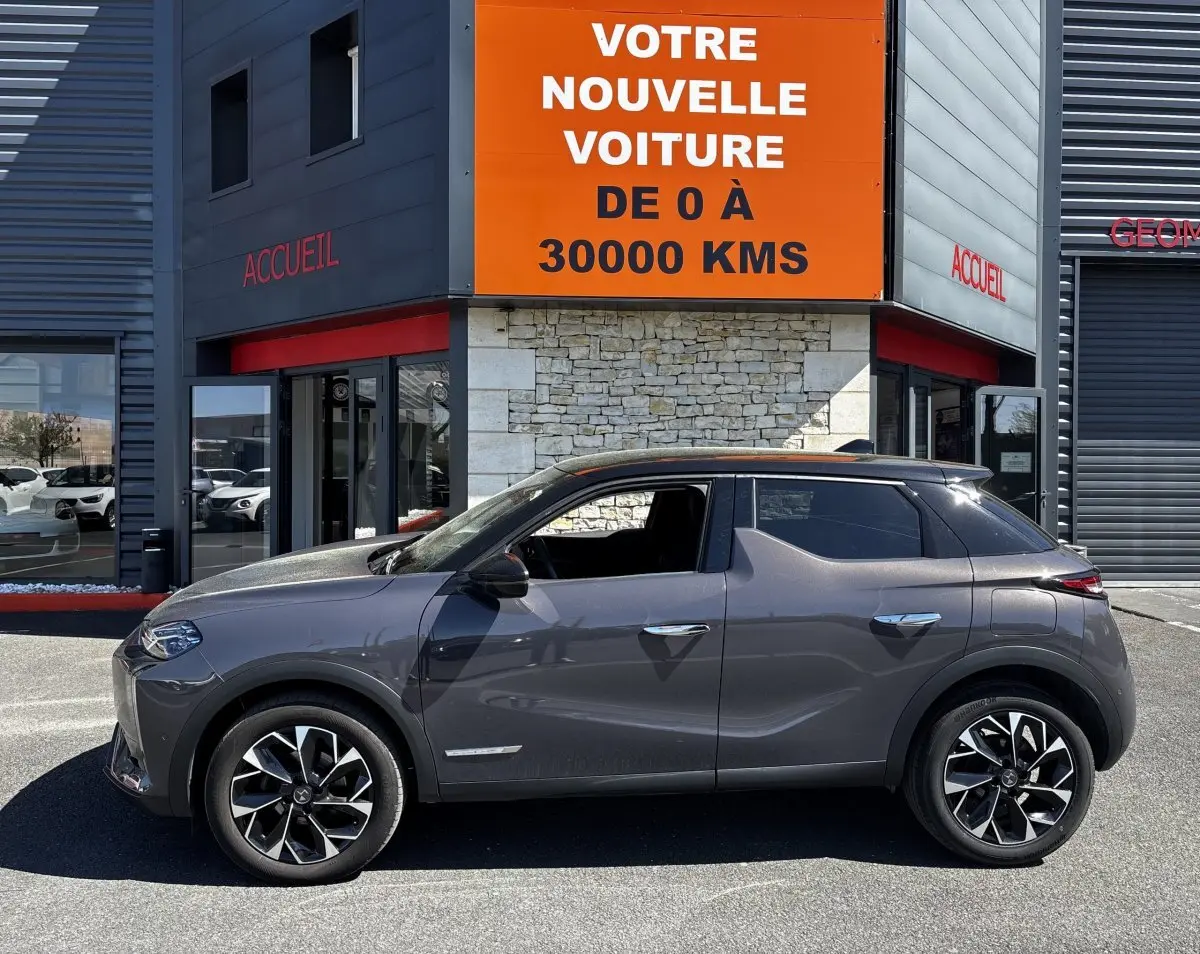 Vue de profil côté gauche d'une DS3 1.2 PureTech 130 gris foncé garée devant un bâtiment avec panneau orange.