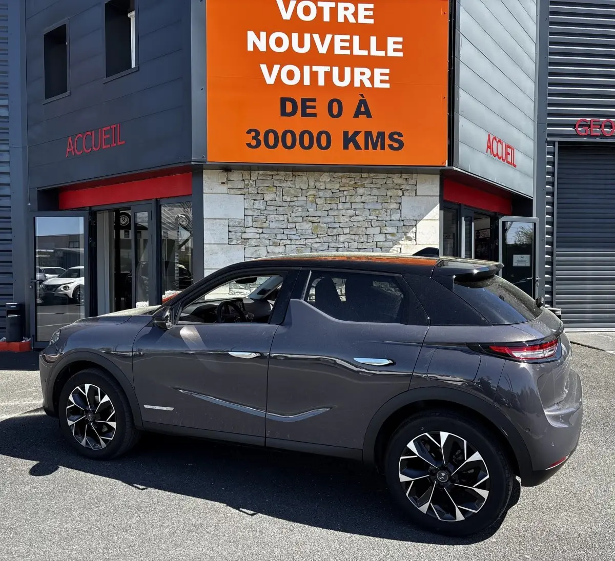 Vue de profil côté gauche d'une DS3 gris foncé garée devant un bâtiment avec panneau orange "Votre nouvelle voiture".
