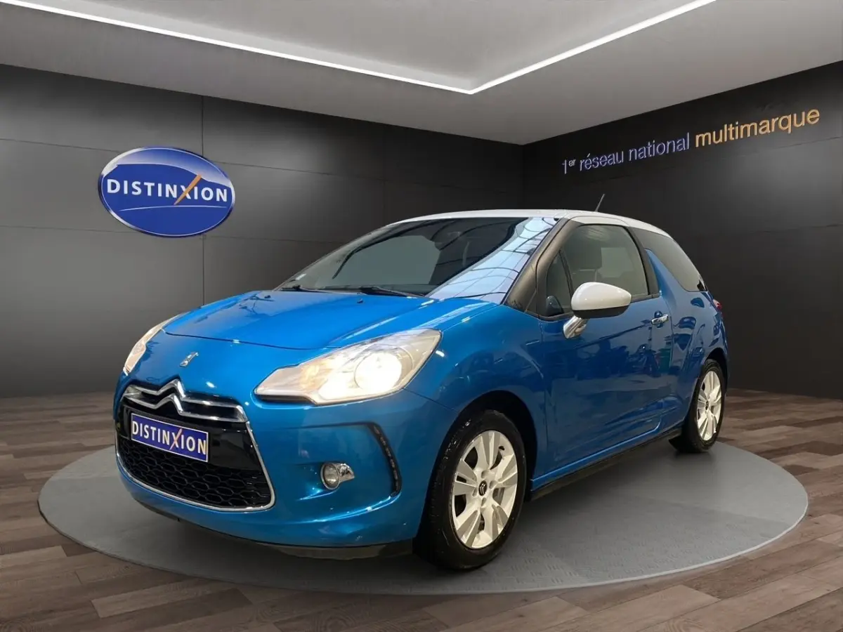 Citroën DS3 e-HDi 90 bleu vue 3/4 avant droit avec toit et rétroviseurs blancs, phares allumés en showroom.