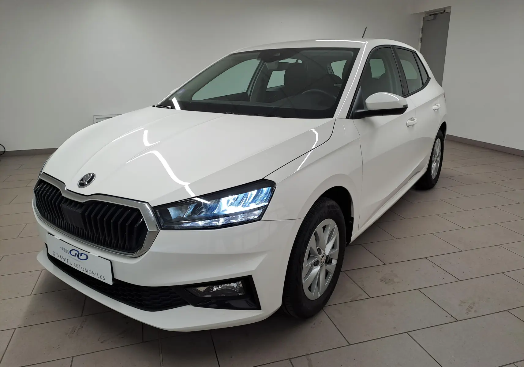 Skoda Fabia blanc candy vue 3/4 avant droit, phares allumés et calandre noire distinctive dans un intérieur neutre.