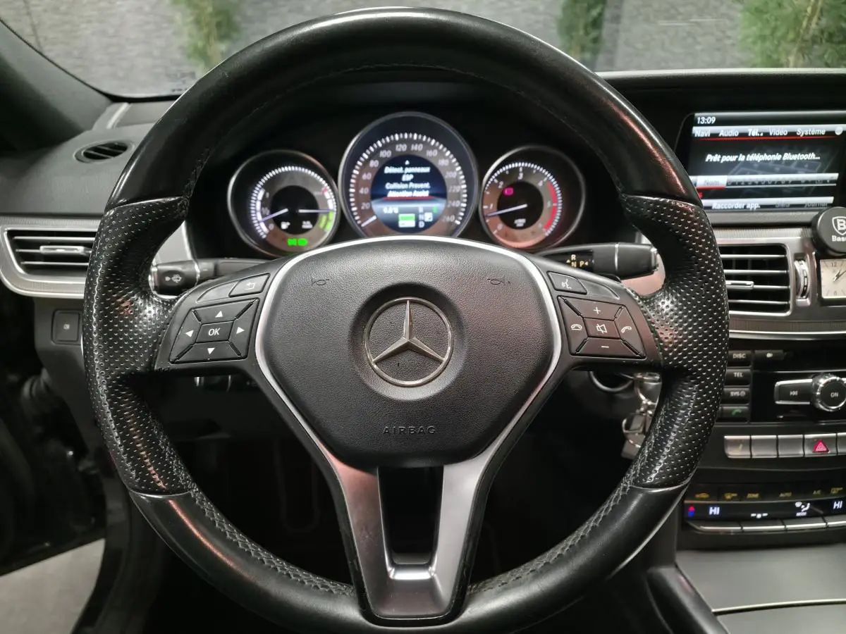 Vue rapprochée du volant en cuir noir perforé de la Mercedes Classe E E 300 BlueTEC Hybrid 2015 avec tableau de bord et écran multimédia.