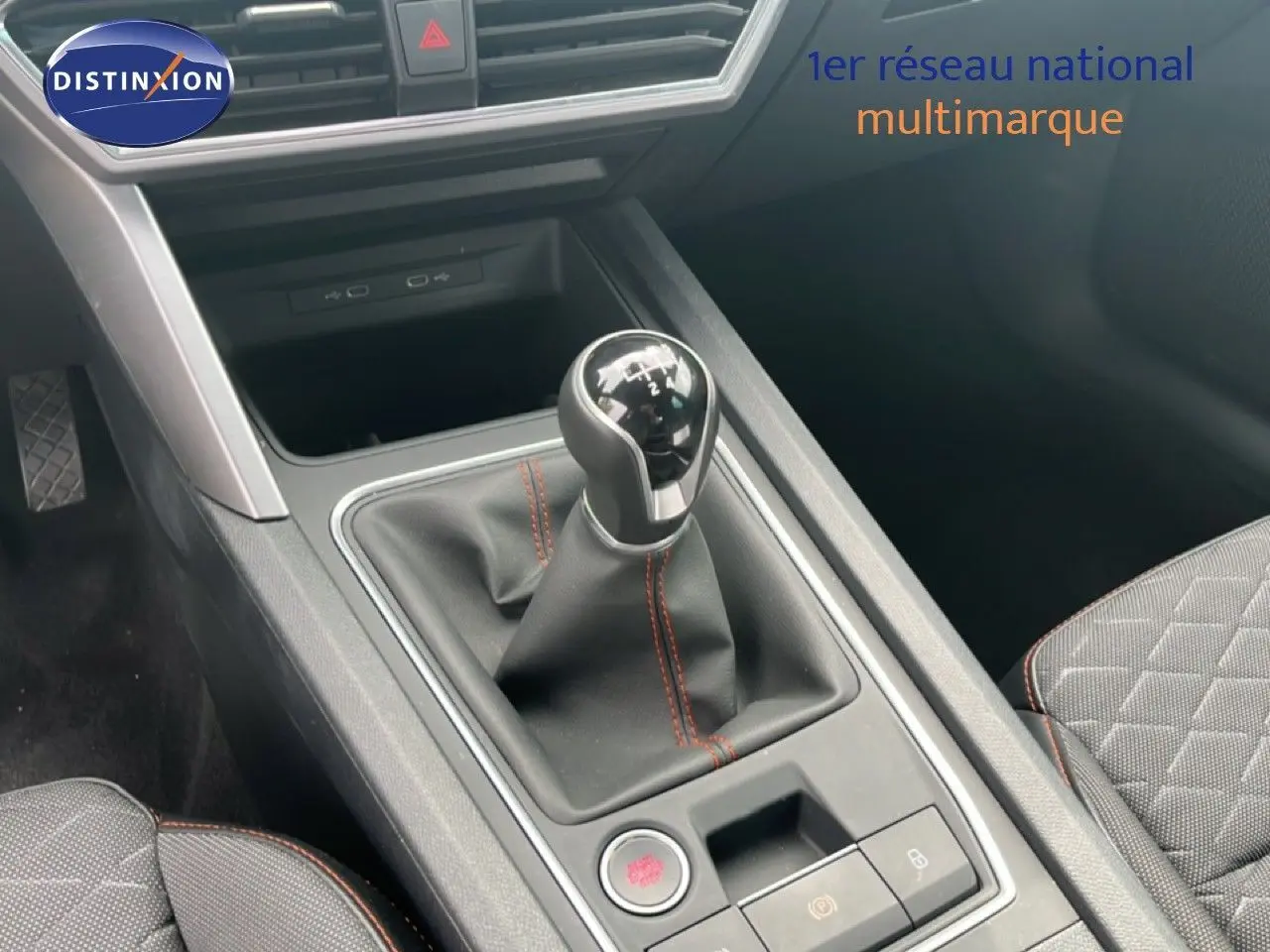 Gros plan sur la console centrale avec levier de vitesse manuel et surpiqûres rouges, siège tissu gris noir visible.