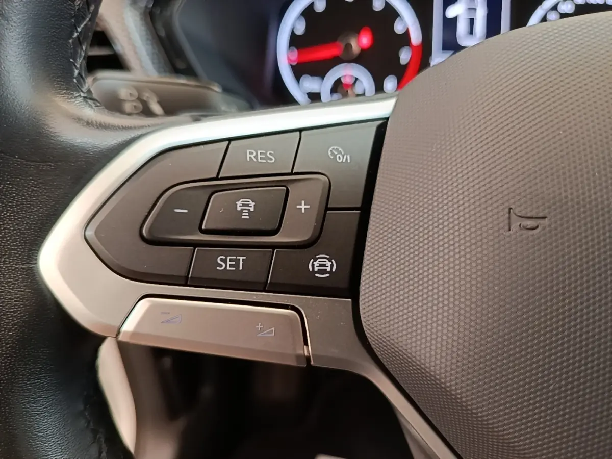 Gros plan sur les commandes de régulateur adaptatif au volant cuir alu du Volkswagen T-Cross blanc, tableau de bord en arrière-plan flou.