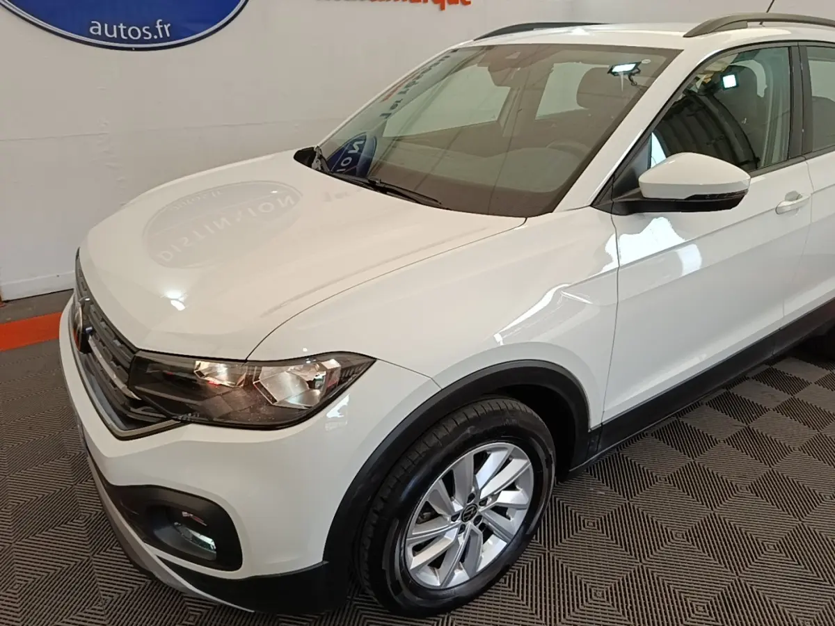 Vue 3/4 avant droit d'un Volkswagen T-Cross blanc avec jantes alliage et barres de toit noires en intérieur showroom.