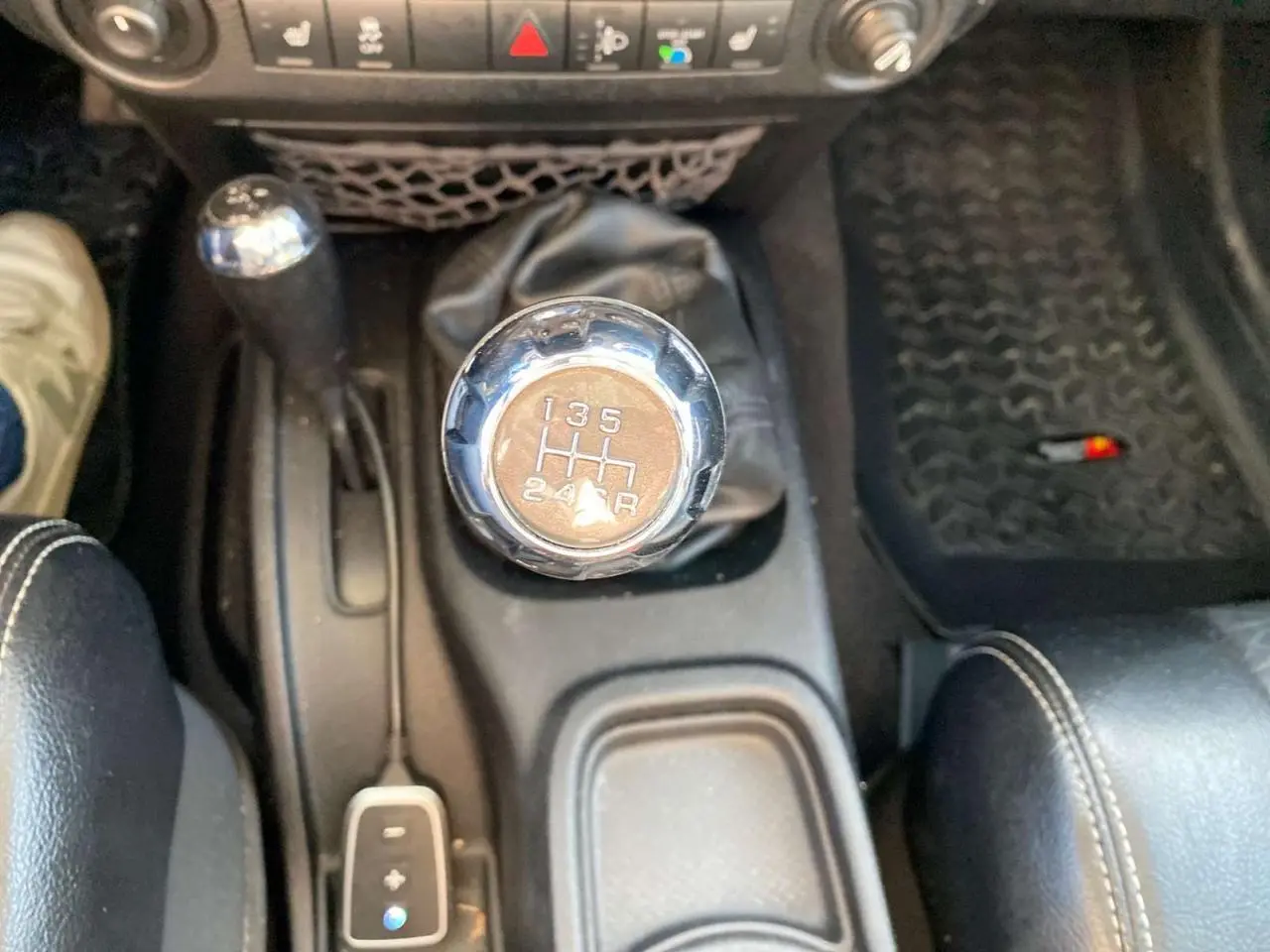 Gros plan sur le levier de vitesses manuel chromé d'une Jeep Wrangler gris clair 2012, avec console et tapis noirs visibles.