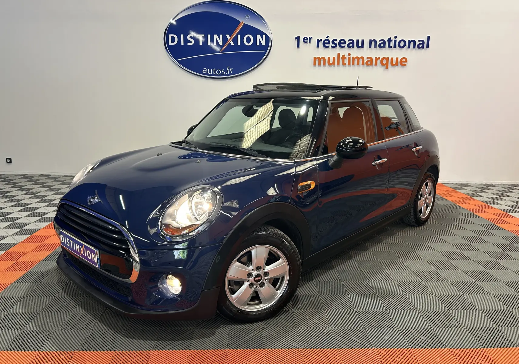 Vue 3/4 avant d'une MINI 5 portes Cooper D bleu foncé avec toit ouvrant, en intérieur sur sol damier gris et orange.