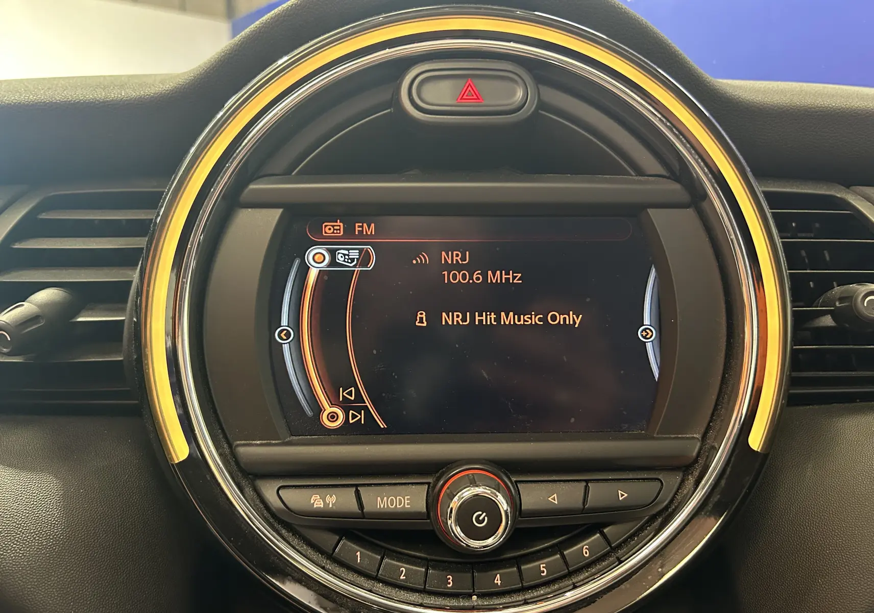 Gros plan sur l’écran central rond du tableau de bord de la MINI 5P, avec éclairage orange et affichage radio NRJ 100.6 MHz.