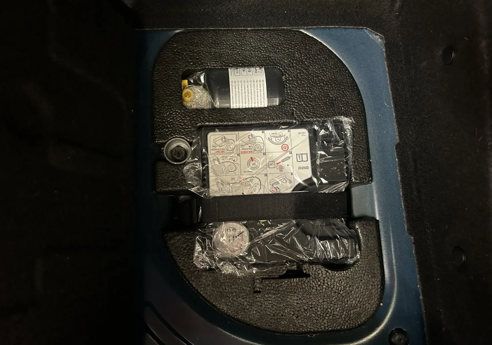 Vue rapprochée du coffre d’une MINI 5 portes bleu, montrant un kit de réparation pneu emballé dans son compartiment.