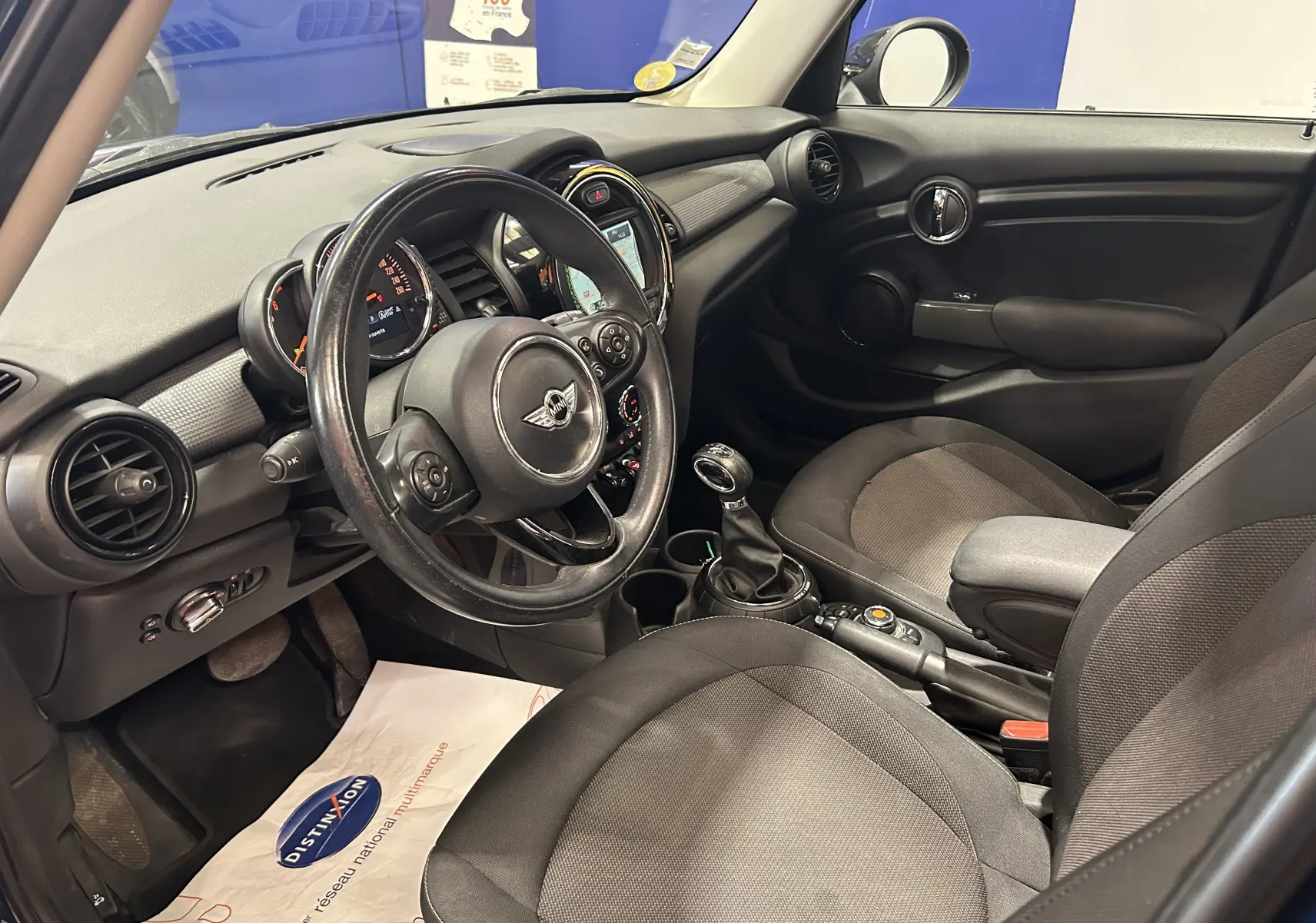 Vue intérieure côté conducteur de la MINI 5P Cooper D 2015, montrant le volant cuir, tableau de bord et sièges tissu gris.