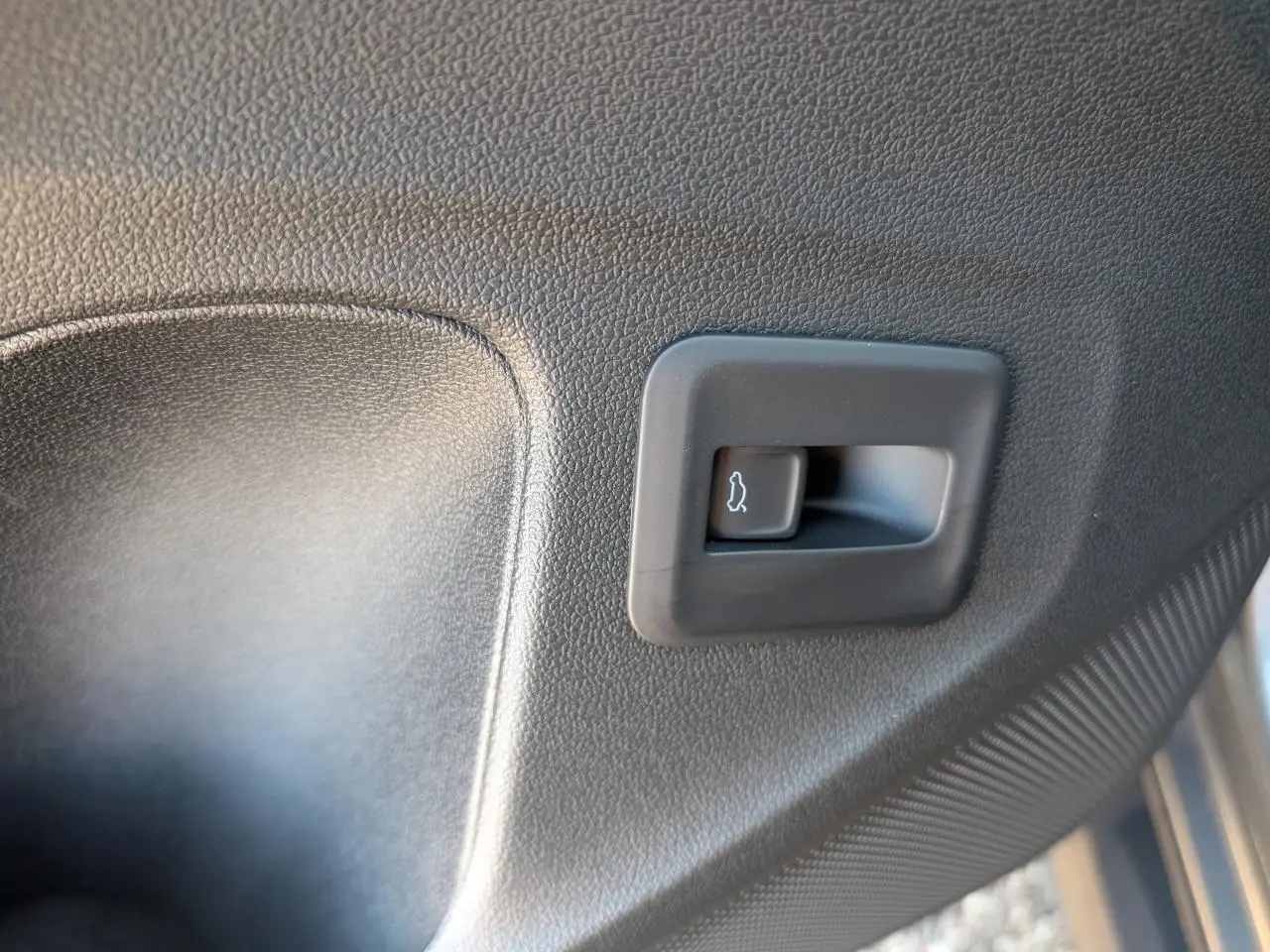 Gros plan sur le bouton d'ouverture électrique du coffre, intégré dans le panneau intérieur noir du Volkswagen T-Roc gris.