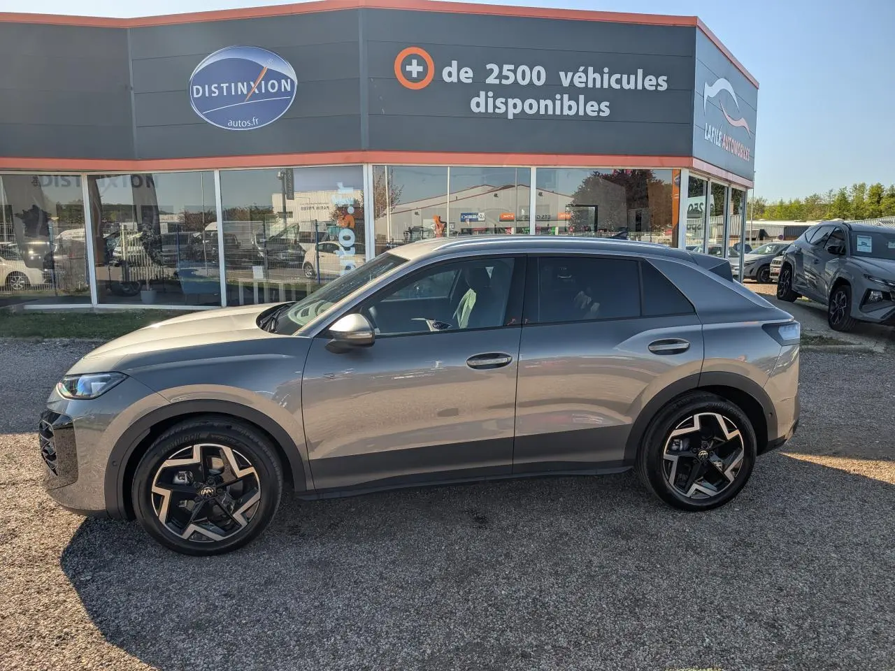 Vue de profil côté gauche d'un Volkswagen T-Roc gris métallisé avec jantes noires et toit noir, stationné devant un showroom.
