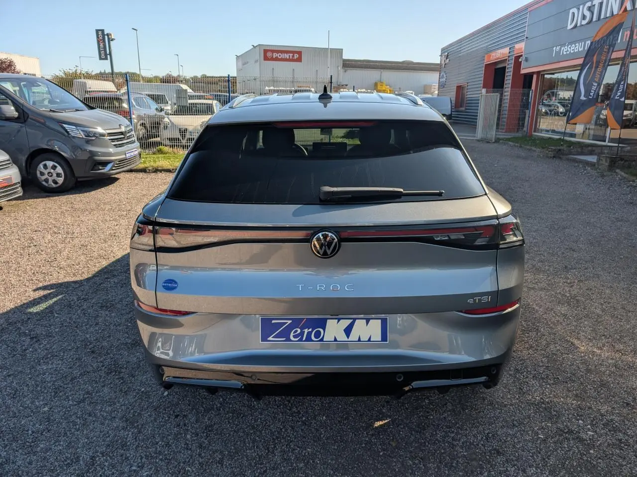 Vue arrière d'un Volkswagen T-Roc gris métallisé avec bandeau lumineux et logo VW rétroéclairé, stationné en extérieur.