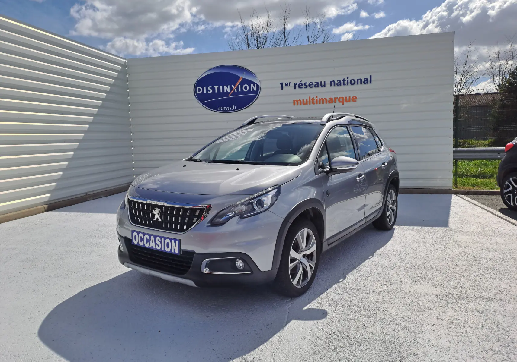 Peugeot 2008 gris en 3/4 avant droit, avec toit ouvrant et jantes alliage, stationné devant un panneau publicitaire.