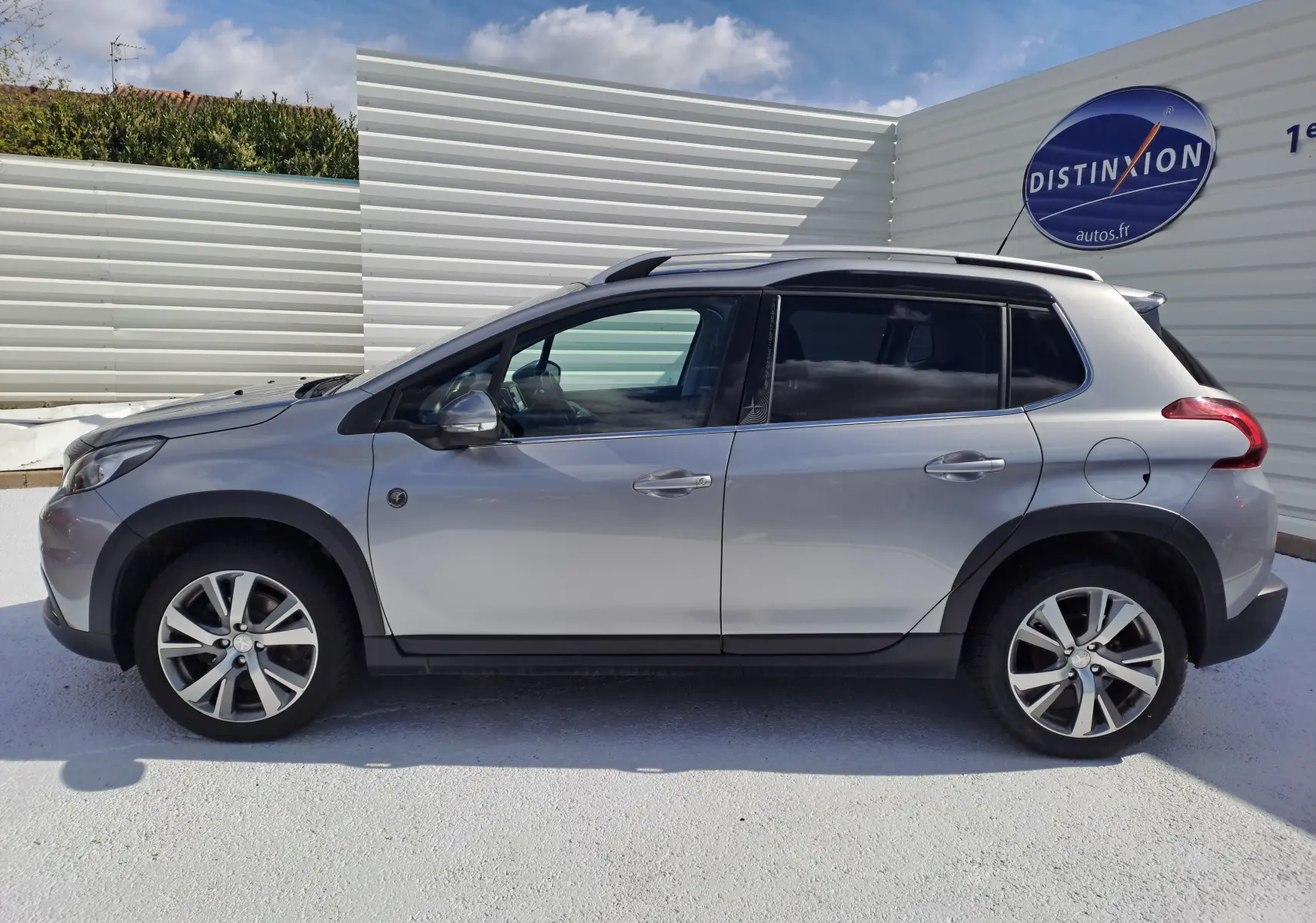 Profil côté gauche d'un Peugeot 2008 gris 2019 avec jantes alliage et barres de toit visibles sur fond clair.