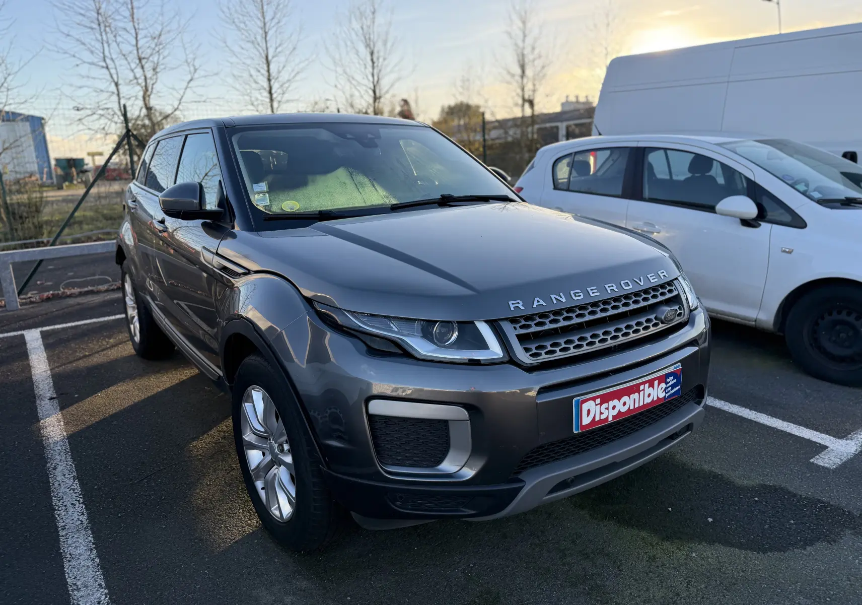 Range Rover Evoque gris vue 3/4 avant droit, avec calandre distinctive et plaque "Disponible" sur parking extérieur.