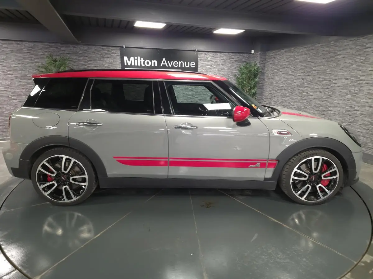 Vue de profil côté gauche d'une MINI Clubman John Cooper Works gris avec toit et détails rouges sportifs en intérieur showroom.