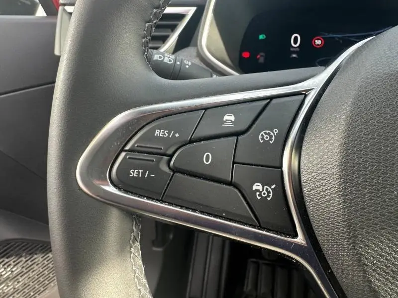 Gros plan sur les commandes au volant gauche de la Renault Clio 2025 Rouge Flamme, avec régulateur de vitesse et aides à la conduite.