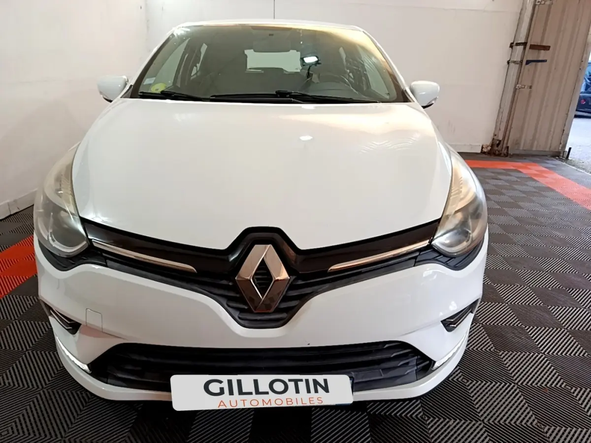 Vue frontale d'une Renault Clio blanche 2016 avec logo Renault proéminent et phares effilés dans un garage.