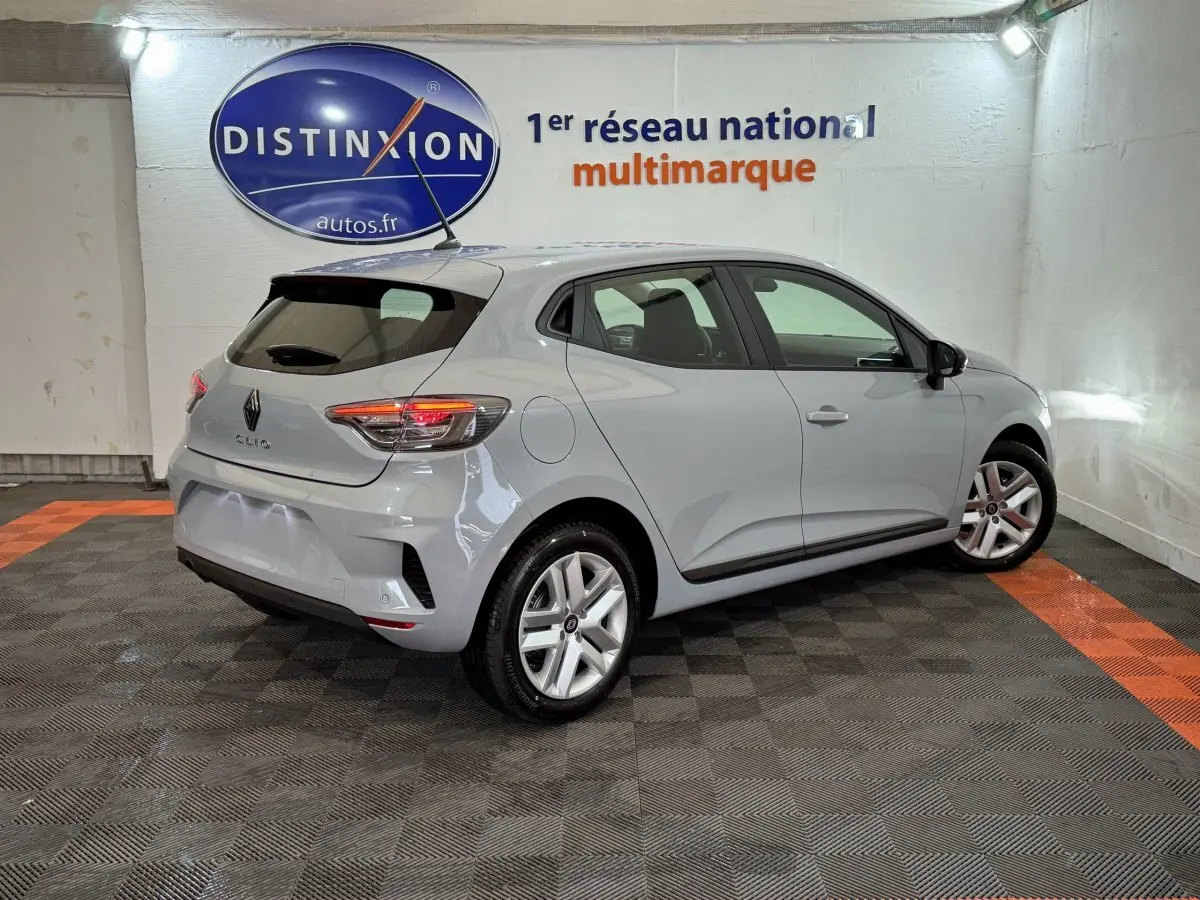 Vue 3/4 arrière droite d'une Renault Clio gris clair 2025 dans un showroom avec feux arrière allumés.