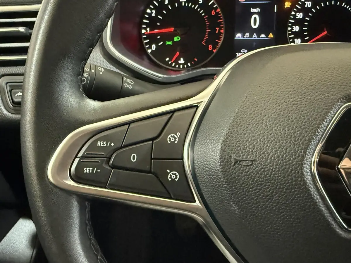 Gros plan sur le volant cuir synthétique noir de la Renault Clio avec commandes de régulateur de vitesse et tableau de bord éclairé.