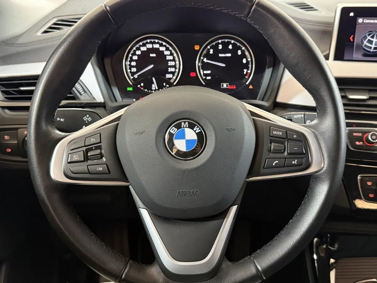 Vue rapprochée du volant cuir noir multifonctions d’une BMW X2 sDrive18i 2023 avec tableau de bord numérique en arrière-plan.