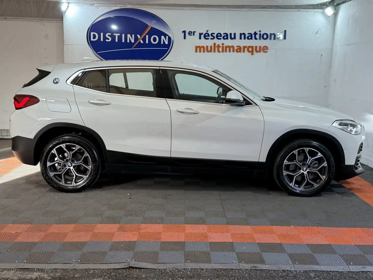 BMW X2 sDrive18i blanc vue de profil côté gauche, avec jantes alliage et feux arrière allumés dans un showroom.