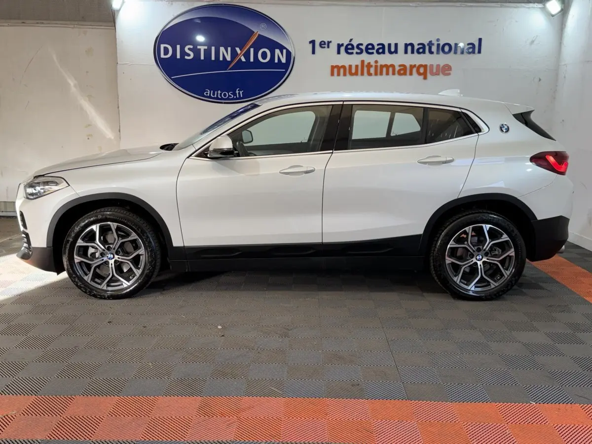 Profil côté gauche d’un BMW X2 sDrive18i Lounge blanc, avec jantes alliage et feux arrière LED visibles en intérieur showroom.