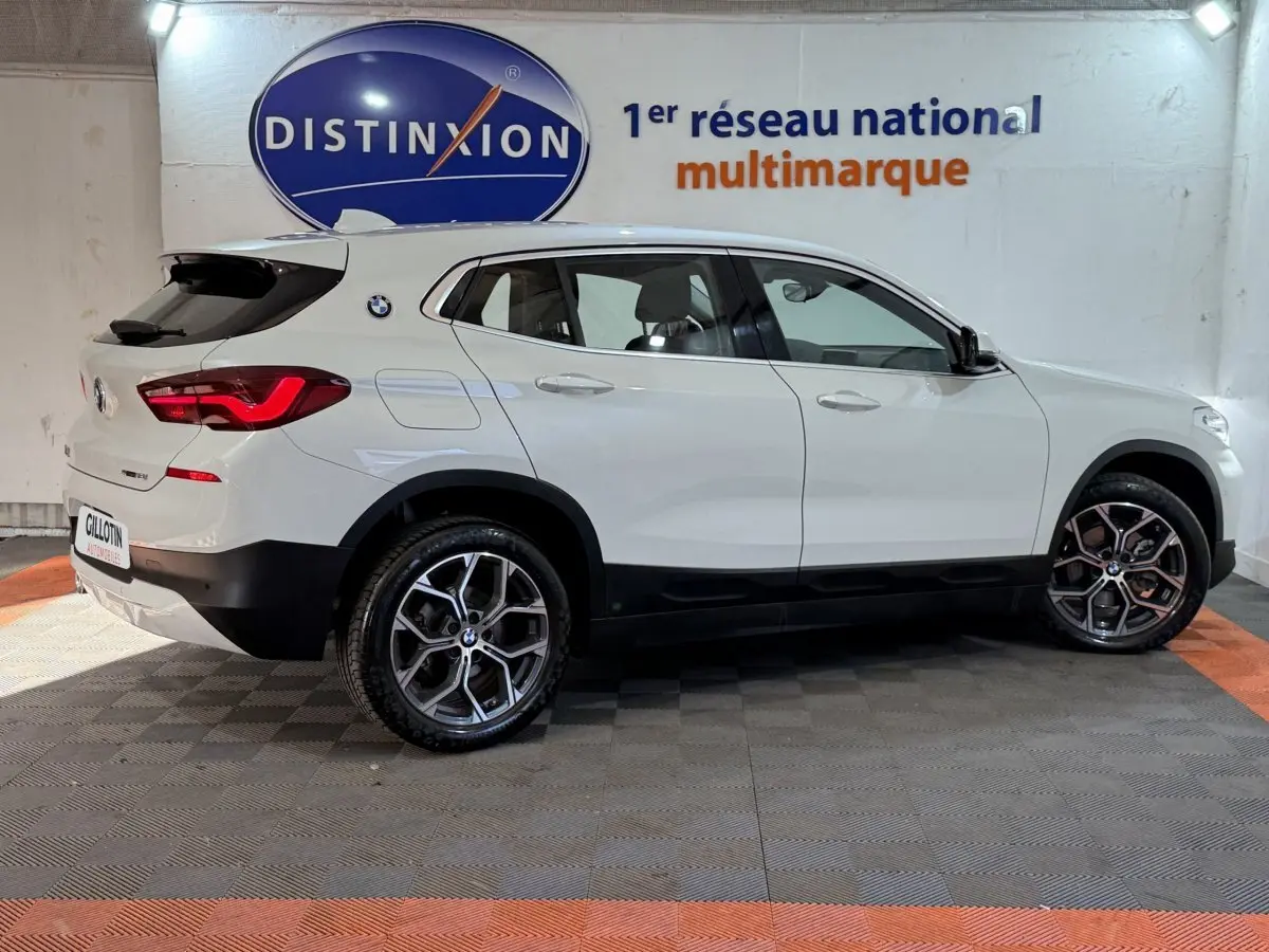 Profil droit d'un BMW X2 sDrive18i Lounge blanc, avec jantes alliage et feux arrière LED visibles en intérieur showroom.
