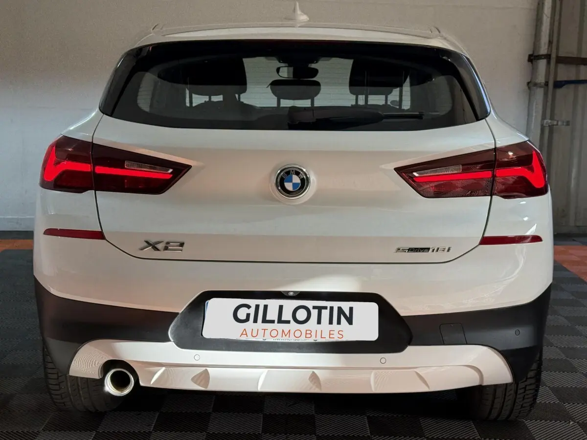 Vue arrière d’un BMW X2 sDrive18i blanc avec feux LED allumés et logo central visible dans un garage.