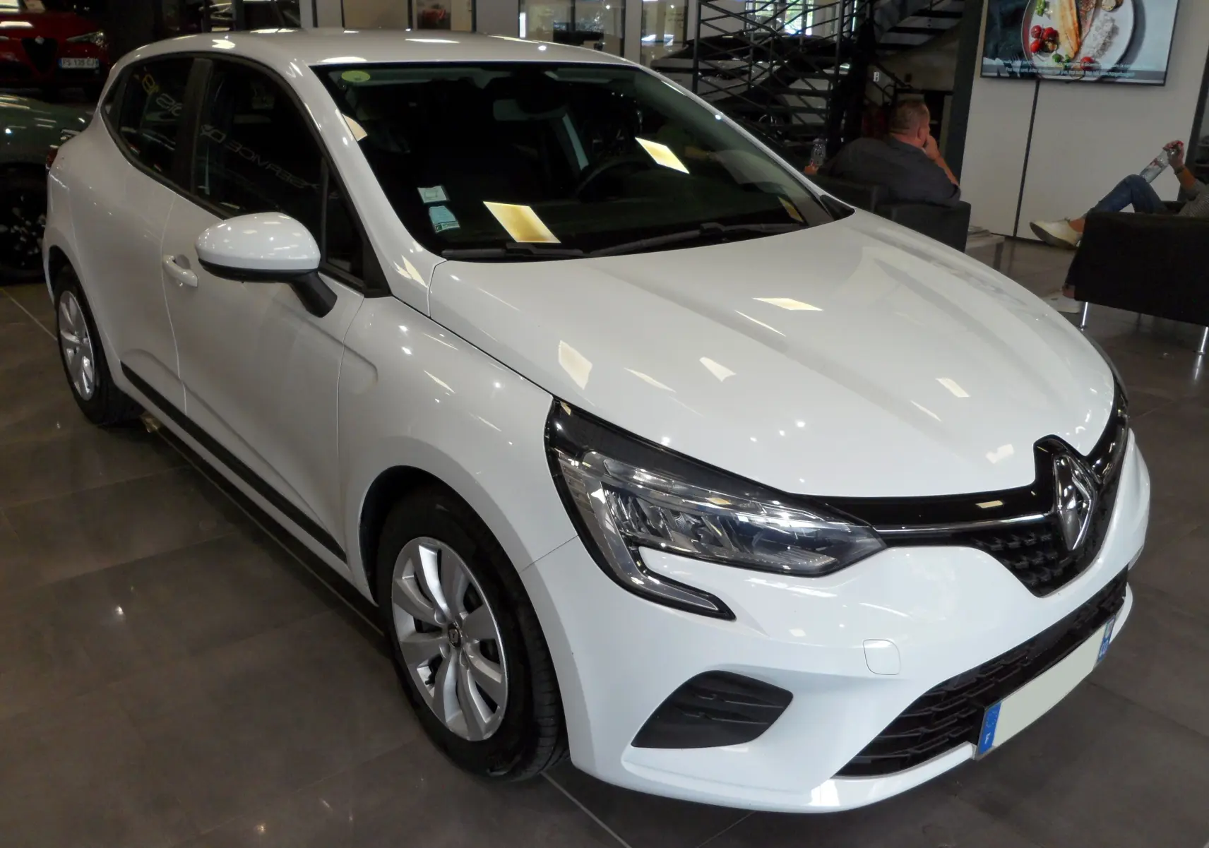 Renault Clio V blanc glacier vue 3/4 avant droit dans un showroom avec jantes alliage et calandre noire.