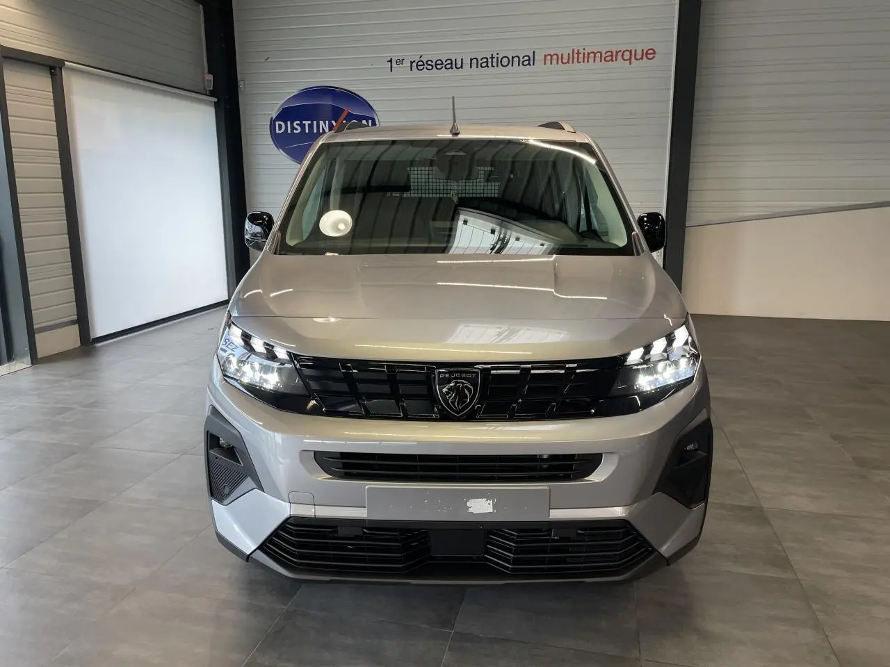 Vue avant du Peugeot Rifter gris Artense avec calandre noire et signature lumineuse LED allumée en intérieur showroom.