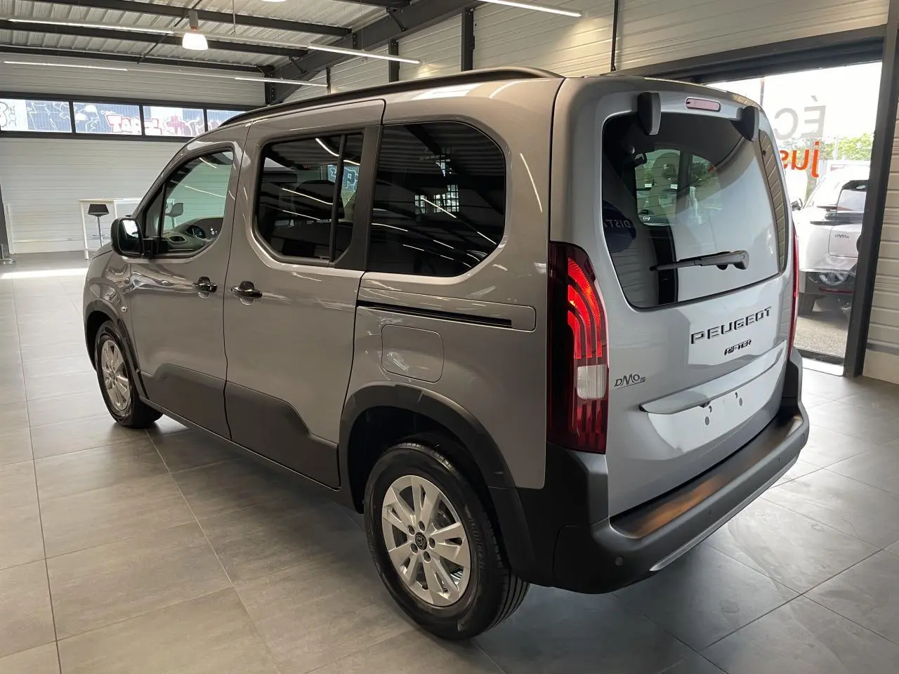 Peugeot Rifter gris Artense vu en 3/4 arrière droit, avec vitres teintées et feux arrière allumés en intérieur showroom.