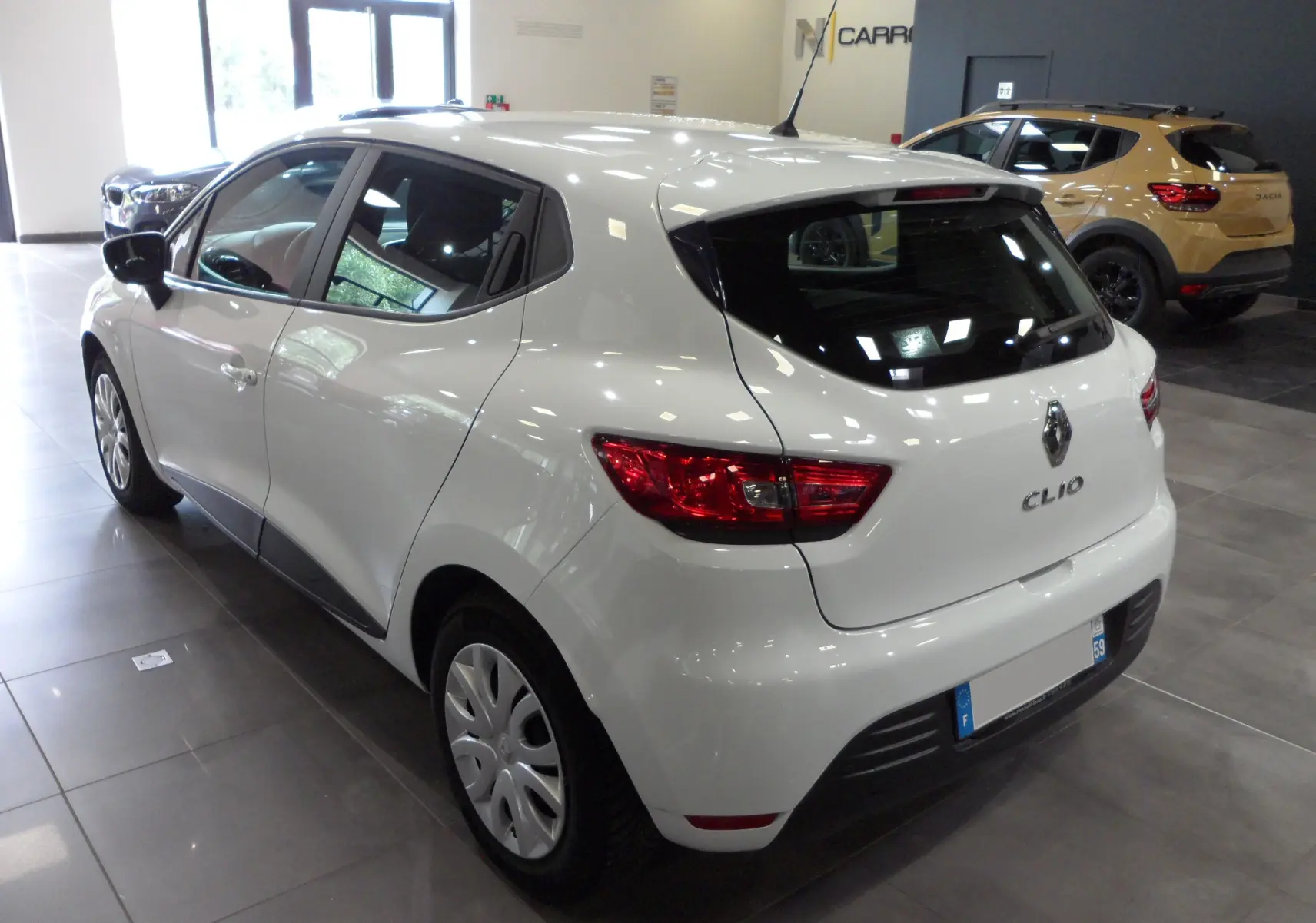 Vue 3/4 arrière droite d'une Renault Clio IV Société blanche Glacier, avec feux arrière rouges et jantes acier en intérieur showroom.