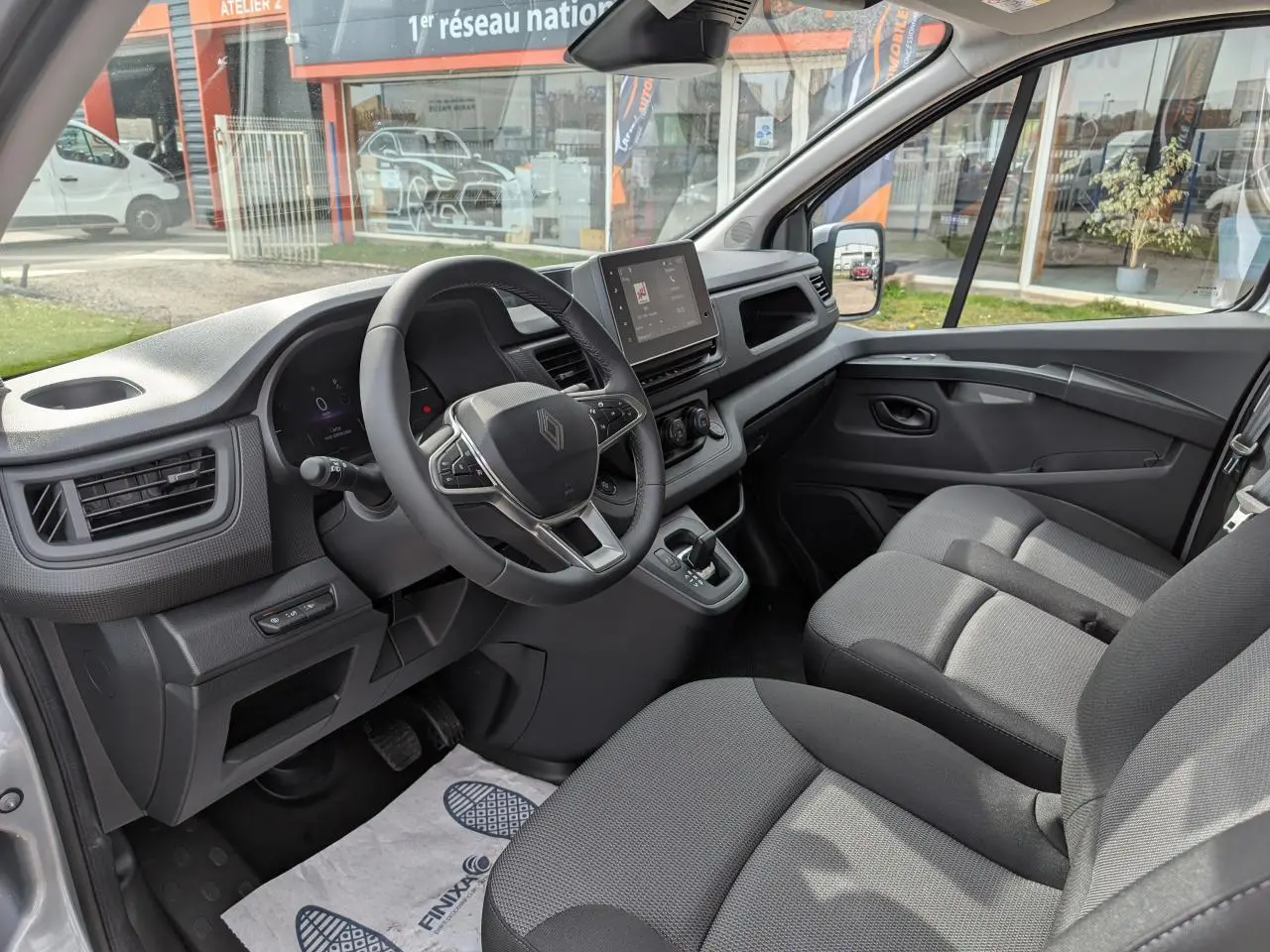 Vue intérieure côté conducteur du Renault Trafic Fourgon gris, montrant le volant, la console centrale et la banquette passager 2 places.
