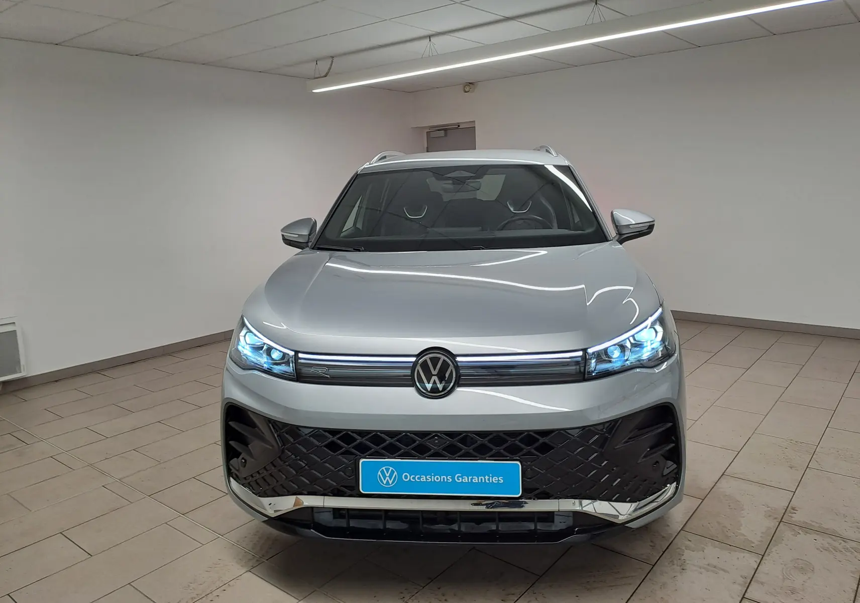Vue avant d'un Volkswagen Tiguan 2.0 TDI 150 DSG7 R-Line en argent Dolomite avec phares LED allumés dans un showroom.