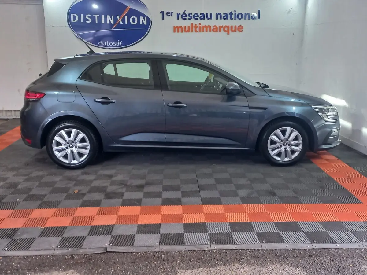 Profil côté gauche d'une Renault Mégane Business TCe 115 gris foncé, stationnée en intérieur sur sol à damiers noir et orange.