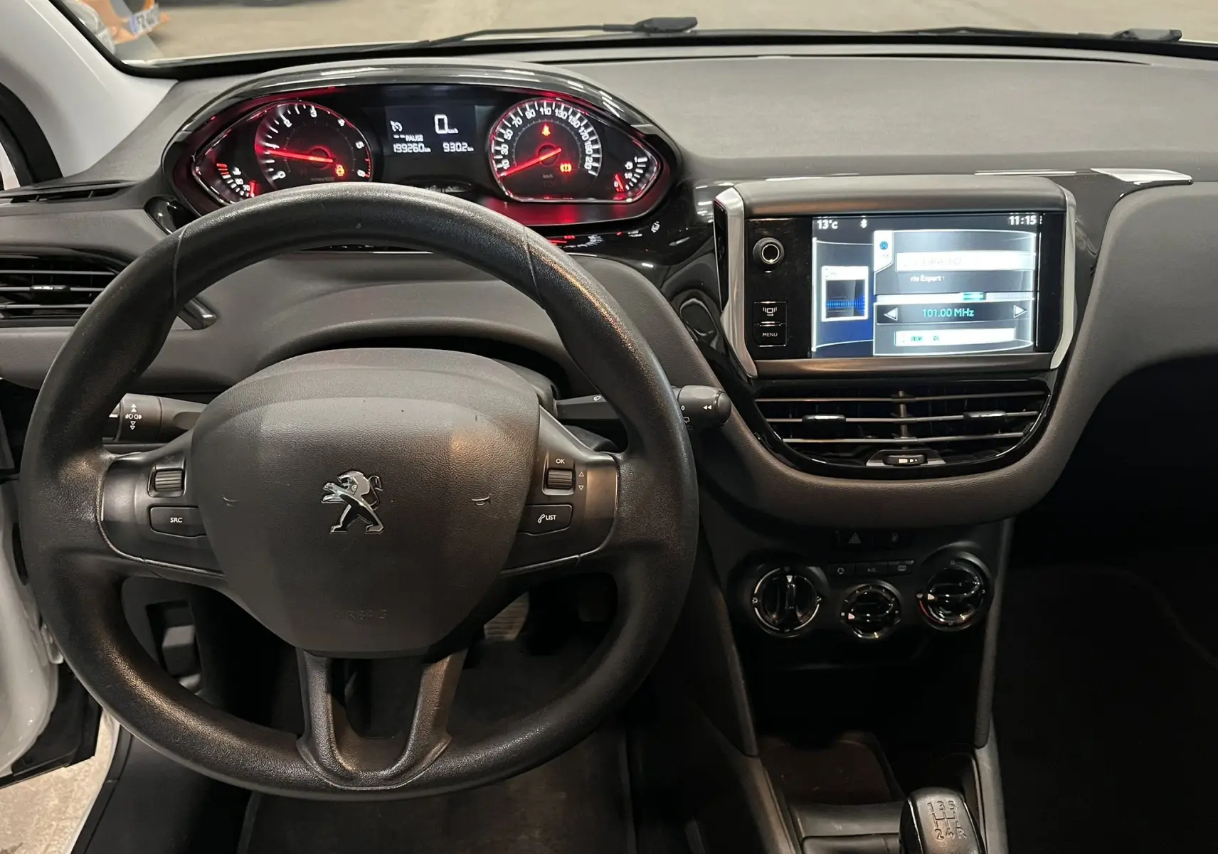 Vue intérieure du tableau de bord et volant noir de la Peugeot 208 blanche, avec écran tactile et compteurs rouges.