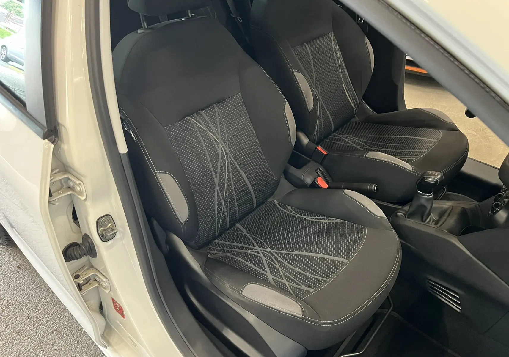 Vue intérieure côté conducteur de la Peugeot 208 blanche, sièges avant en tissu noir avec motifs gris et levier de vitesse manuel.