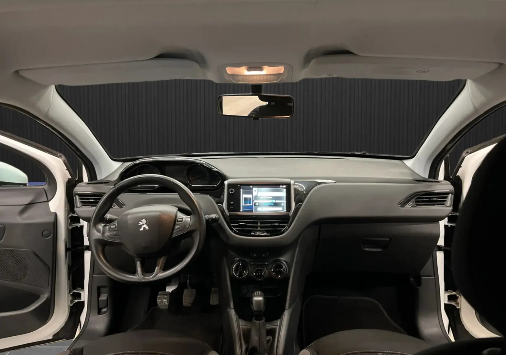 Vue intérieure avant du tableau de bord noir et volant multifonction d'une Peugeot 208 blanche avec portes ouvertes.