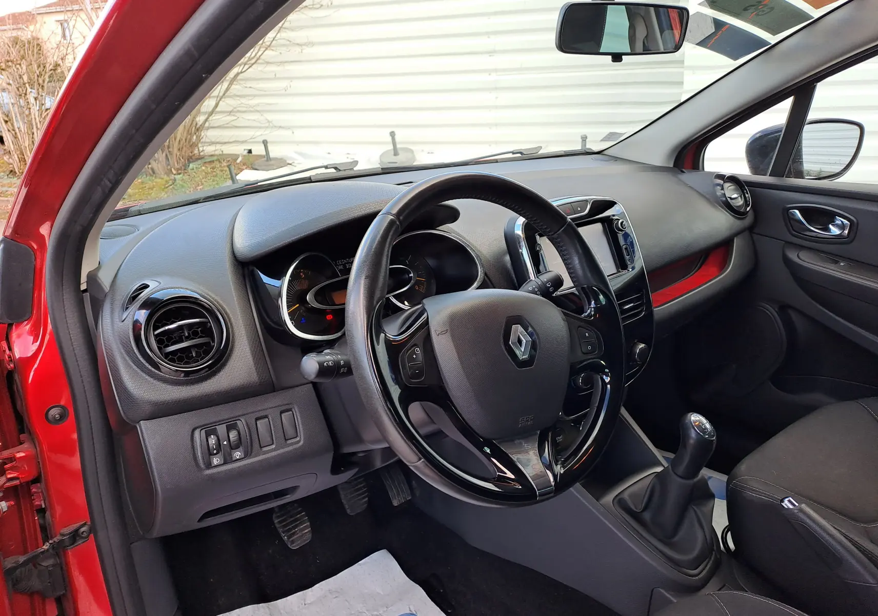 Vue intérieure côté conducteur de la Renault Clio IV rouge, montrant le volant, tableau de bord et levier de vitesses manuel.
