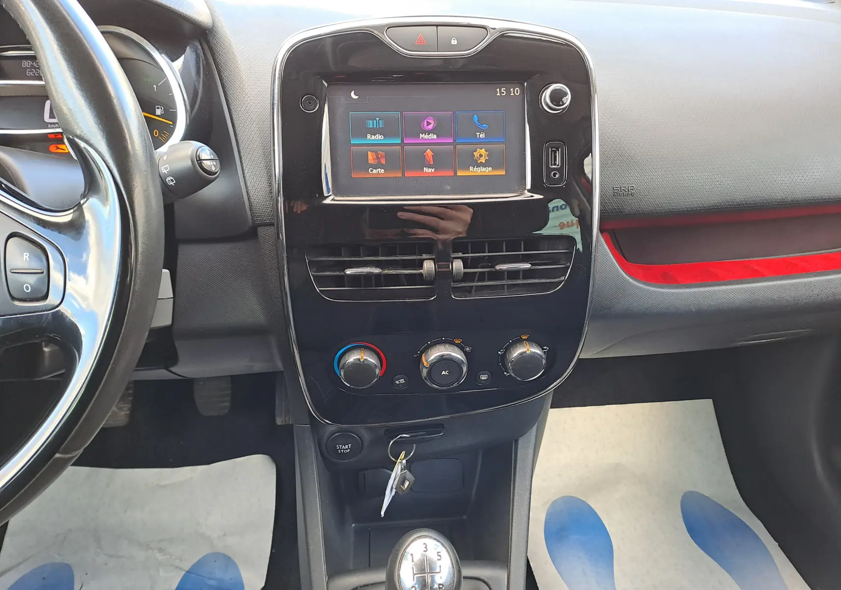 Vue intérieure centrée sur la console centrale noire brillante avec écran tactile et levier de vitesse manuel d'une Renault Clio IV rouge.
