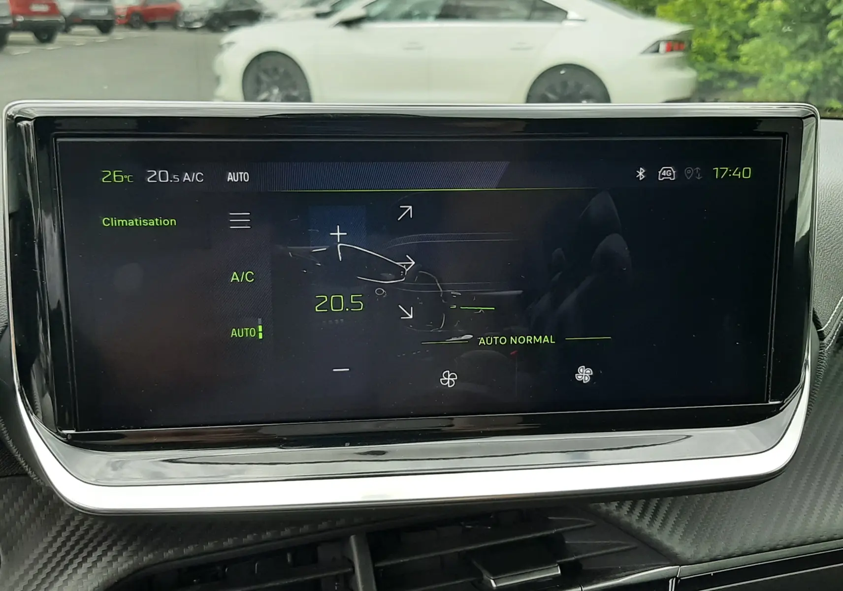 Vue intérieure centrée sur l'écran tactile de climatisation de la Peugeot 208, avec une voiture blanche en arrière-plan flou.