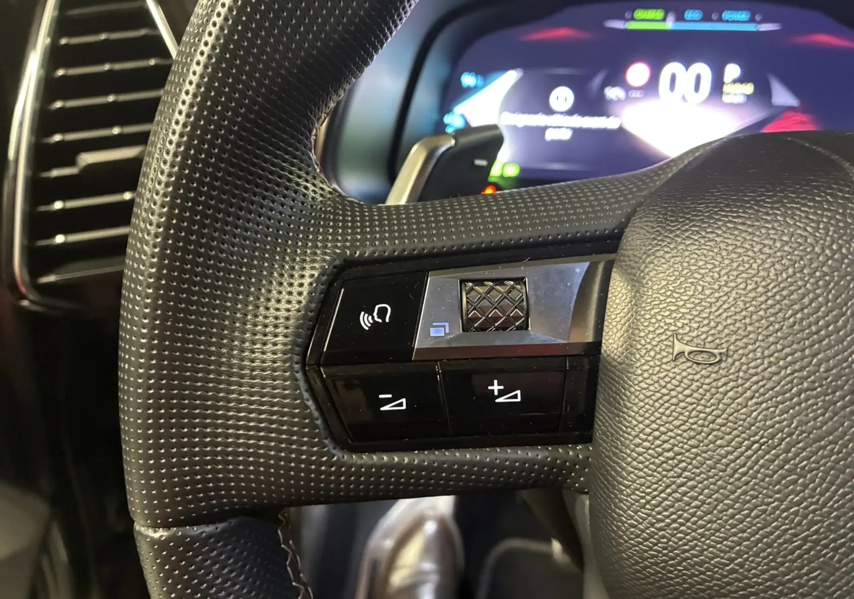 Gros plan sur la partie gauche du volant noir perforé du DS7 E-TENSE 2024 avec commandes intégrées et tableau de bord numérique flou.